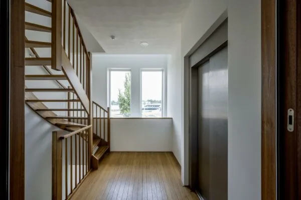 Licht trappenhuis aan de De Ruijterkade met houten trap, lift en drie hoge ramen.